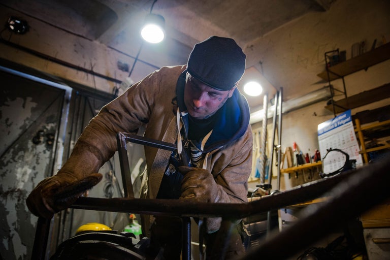 Ein Mann mit Arbeitskleidung und Handschuhen arbeitet an einer Metallkonstruktion in einer Werkstatt.