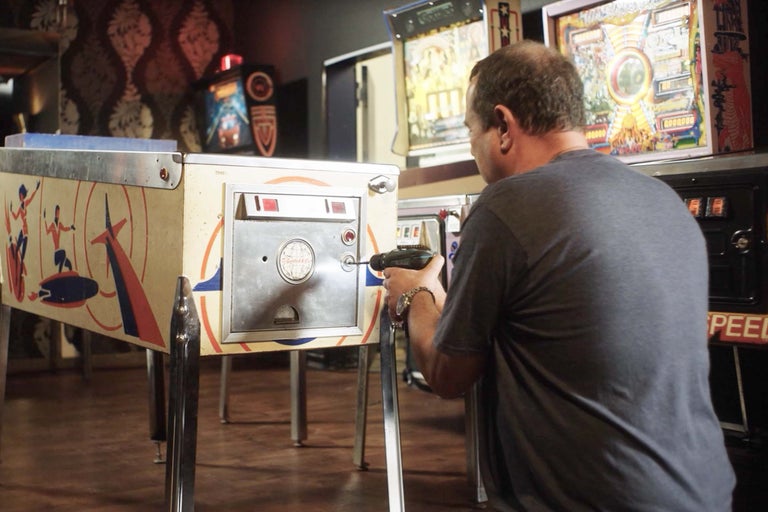 Ein Mann repariert einen Flipperautomaten mit einem Akkuschrauber in einer Spielhalle