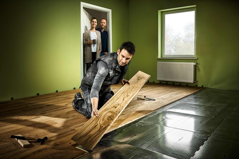 Ein Mann verlegt Laminatboden in einem Raum mit Fenster und Heizkörper. Im Hintergrund stehen zwei Personen in einer offenen Tür.