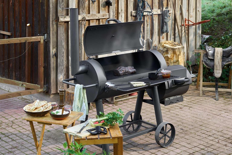 Schwarzer Smoker Grill mit Grillgut und Beilagen im Garten.