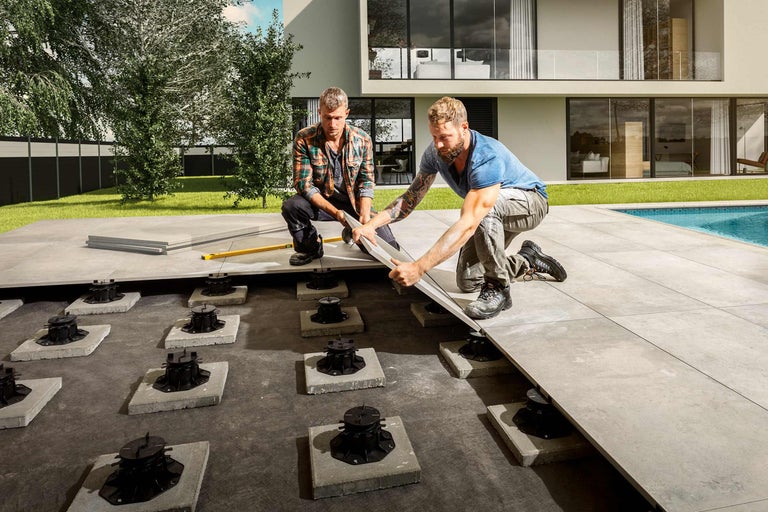 Zwei Handwerker verlegen Terrassenplatten auf Stelzlager in einem Garten.