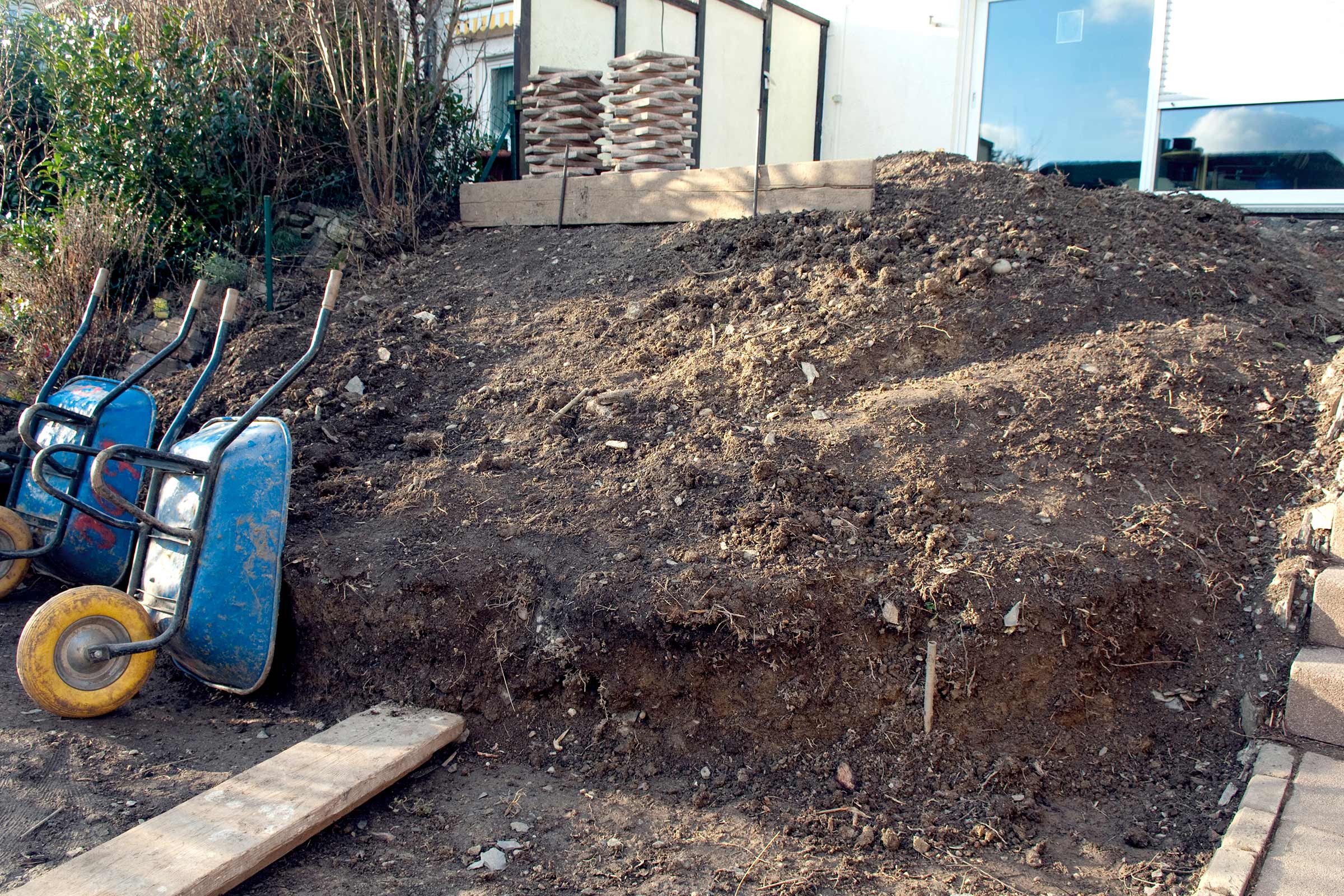 Szene einer Gartengestaltung mit Erdhügeln, zwei Schubkarren und Holzmaterialien.