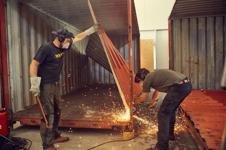 Zwei Arbeiter beim Schneiden von Metall in einem Lagercontainer mit einem Winkelschleifer, wobei Funken sprühen.