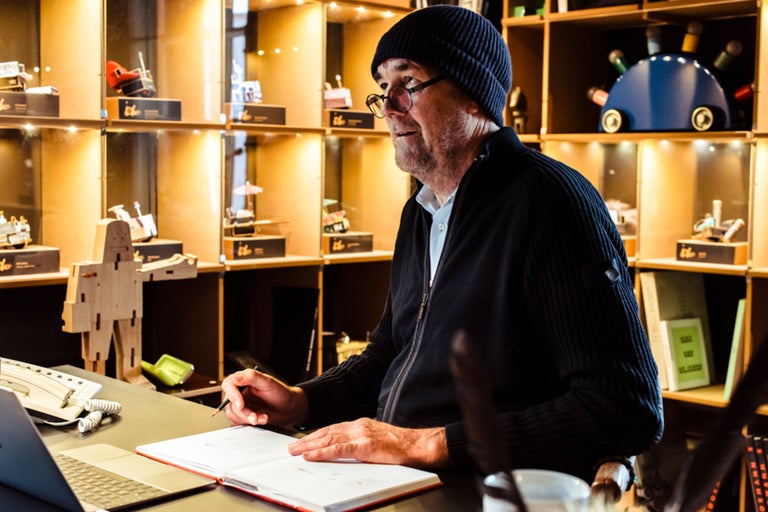 Ein Mann sitzt an einem Schreibtisch mit einem Notizbuch und einem Stift, im Hintergrund Regale mit verschiedenen Gegenständen.