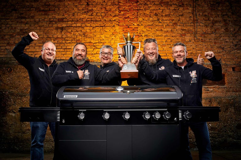 Fünf Männer präsentieren einen schwarzen Gasgrill und einen Pokal vor einer Ziegelwand