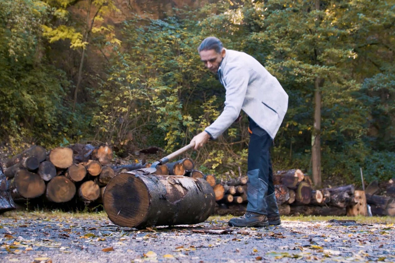 Mann spaltet Holz mit einer Axt vor einem Wald