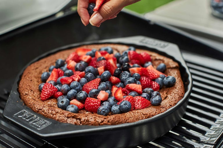 Ein dunkler Kuchen in einer Grillform mit Erdbeeren und Blaubeeren wird von einer Hand dekoriert.
