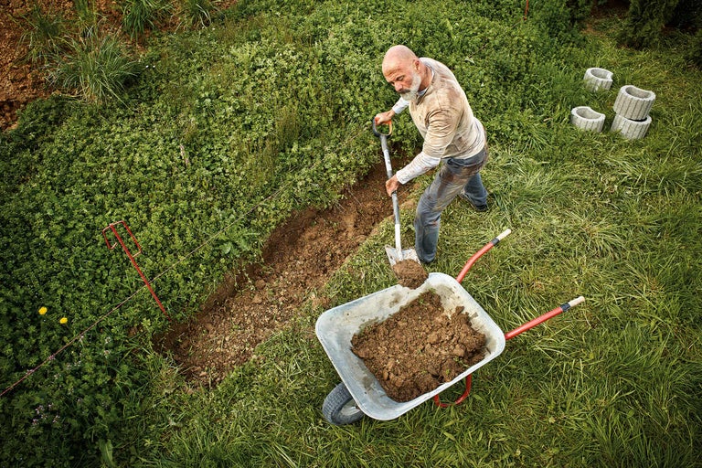 Mann gräbt mit Spaten einen Graben und befüllt eine Schubkarre in einem Garten.