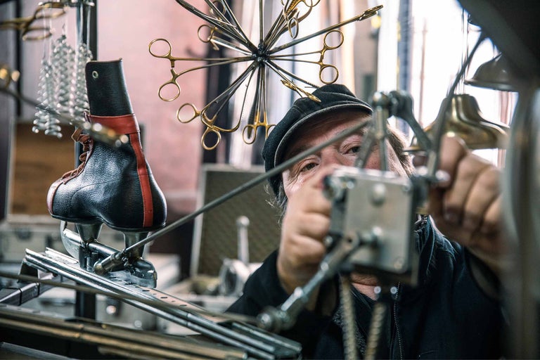 Ein Mann mit Mütze repariert Eiskunstlaufschuhe in einer Werkstatt, umgeben von Werkzeugen.