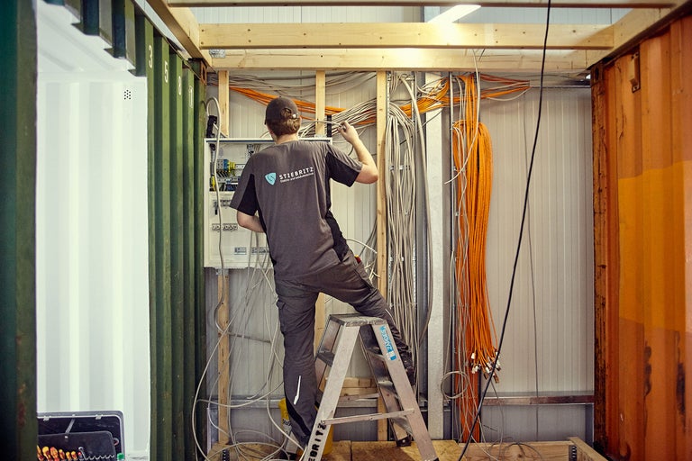 Elektriker auf einer Leiter, der elektrische Leitungen in einem Container installiert
