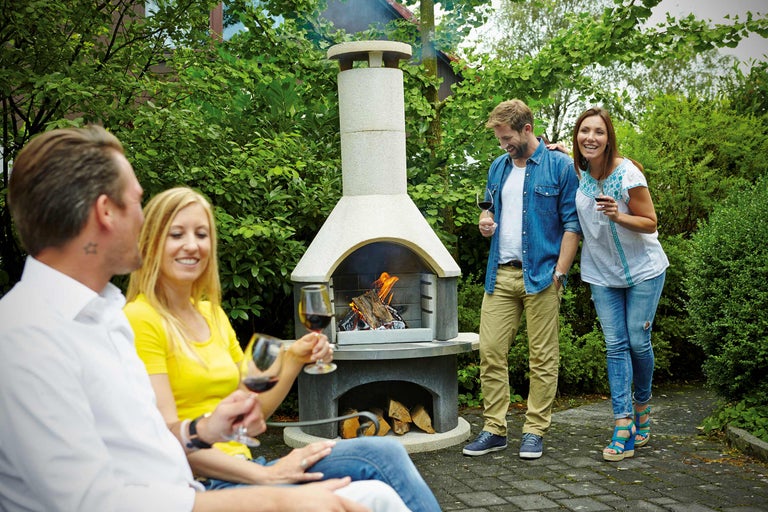 Szene mit Gartengrillkamin, auf dem Holz brennt, und vier Personen, die im Garten mit Weingläsern entspannen.