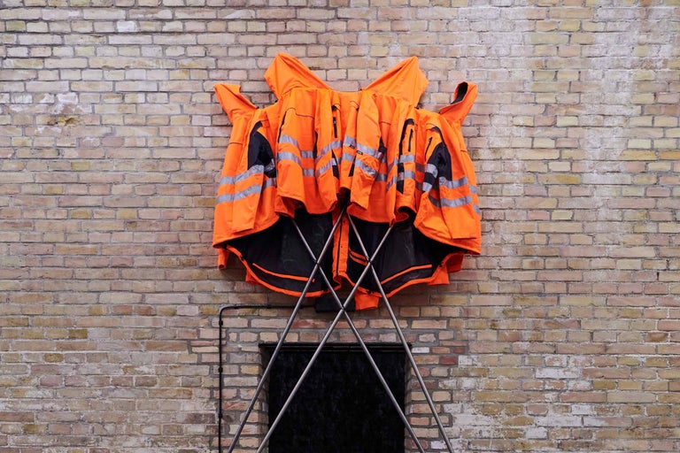 Mehrere Warnwesten mit Reflektorstreifen hängen auf einem Kleiderständer vor einer Backsteinmauer.