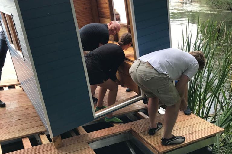 Personen beim Aufbau eines Gartenhauses auf einer Holzterrasse am Wasser.