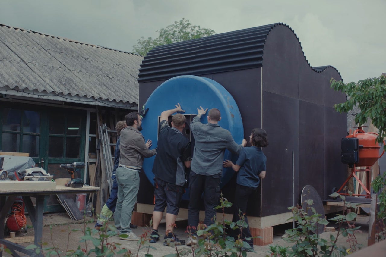 Eine Gruppe von Personen baut ein schwarzes, modernes Gartenhaus mit einer markanten runden Öffnung an der Außenseite auf.