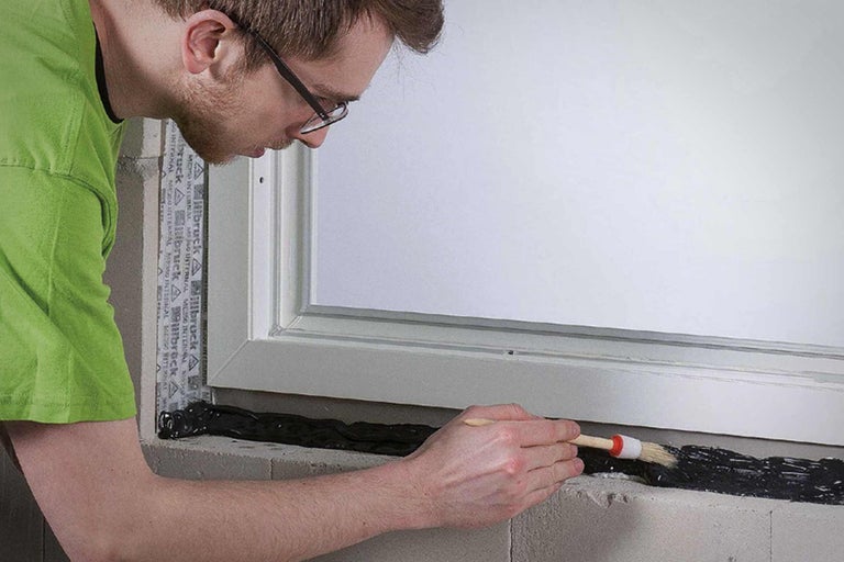 Eine Person trägt mit einem Pinsel eine Dichtungsmasse auf ein Fenster auf.