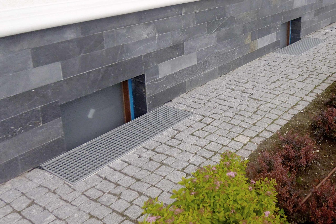 Außenansicht eines Hauses mit Steinfassade, gepflastertem Weg und Entwässerungsrinne vor den Fenstern