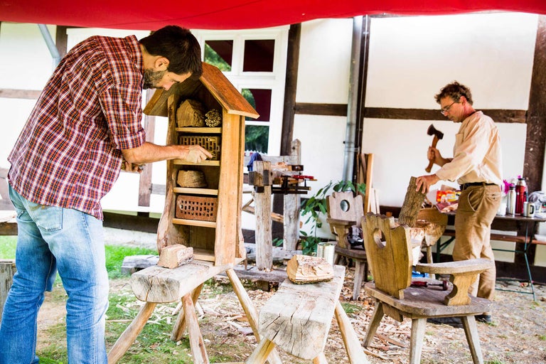 Mann befüllt ein Insektenhotel mit Nistmaterial, während ein anderer Mann im Hintergrund Holz hackt