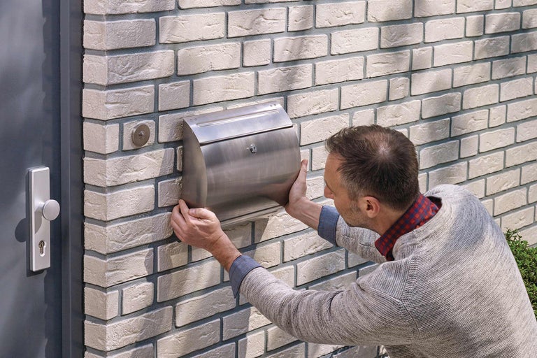 Mann montiert einen Briefkasten aus Stahl an einer Ziegelwand neben einer Haustür.