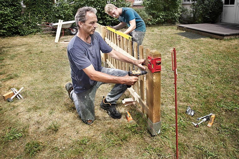 Zwei Männer bauen einen Holzzaun im Garten mit Akkuschrauber und Wasserwaage.