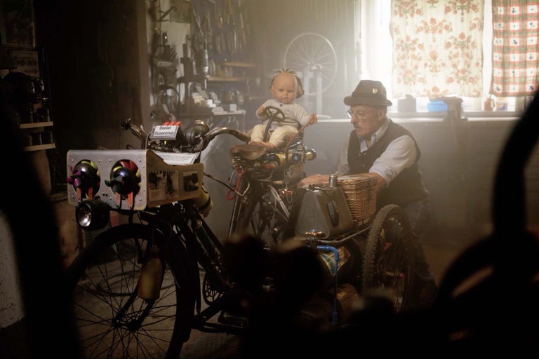 Ein Mann mit Hut und Brille steht neben einem modifizierten Fahrrad mit einem Korb und einer Puppe als Fahrer in einer Werkstatt.