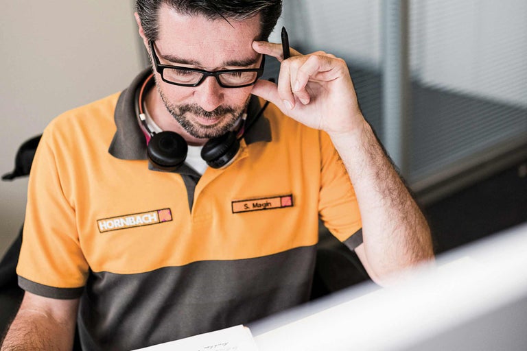 Ein Mitarbeiter von Hornbach denkt nach, mit einem Stift am Kopf und Kopfhörern um den Hals.