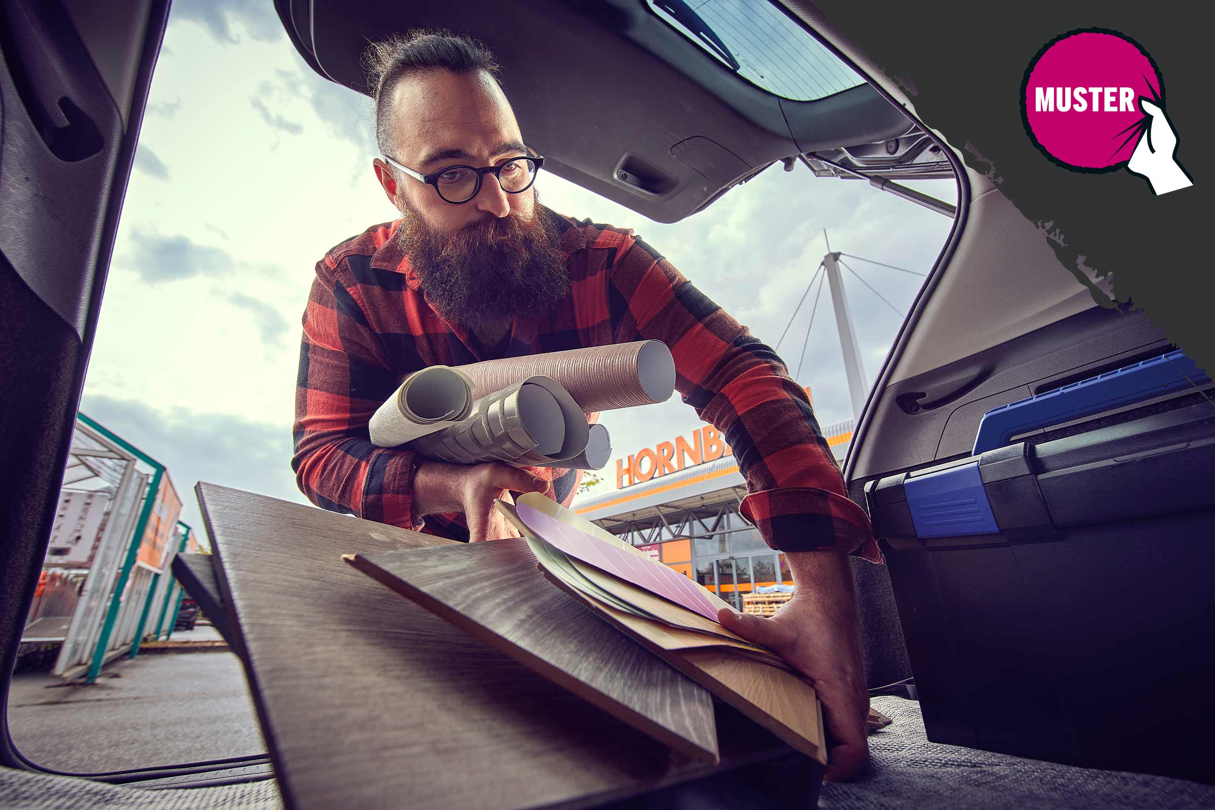 Ein Mann mit Bart belädt den Kofferraum eines Autos mit Laminat, Tapetenrollen und Farbmustern vor einem Hornbach Markt.