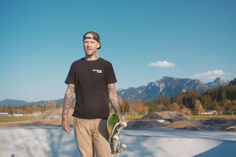 Ein Mann mit Tätowierungen hält ein Skateboard in einem Skatepark.