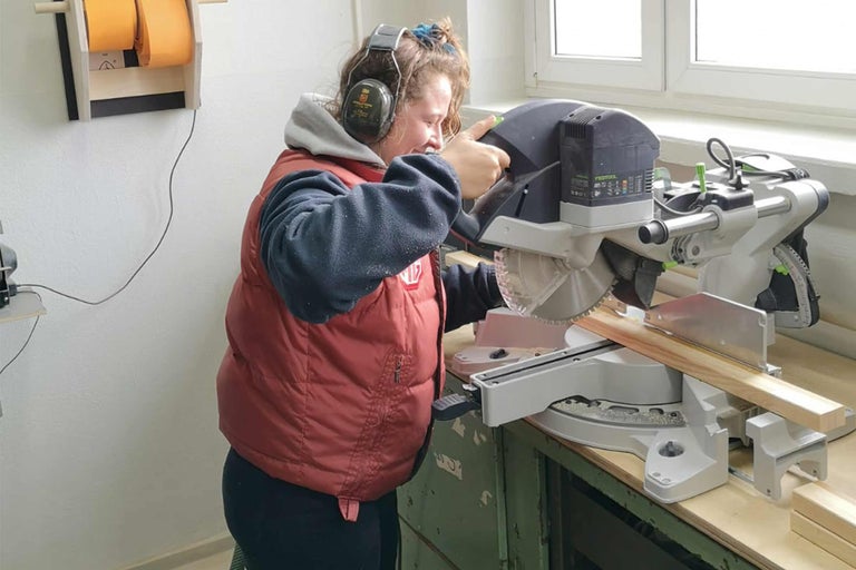 Eine Frau mit Gehörschutz sägt mit einer Kappsäge ein Holzbrett.