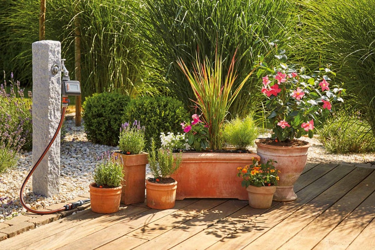 Gartenbewässerungssystem mit Wasserhahn, Schlauch und verschiedenen Blumen in Tontöpfen auf einer Holzterrasse