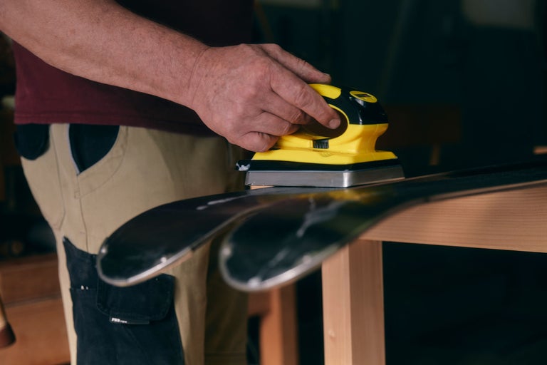 Eine Person schleift Ski mit einem elektrischen Schleifgerät auf einer Werkbank.