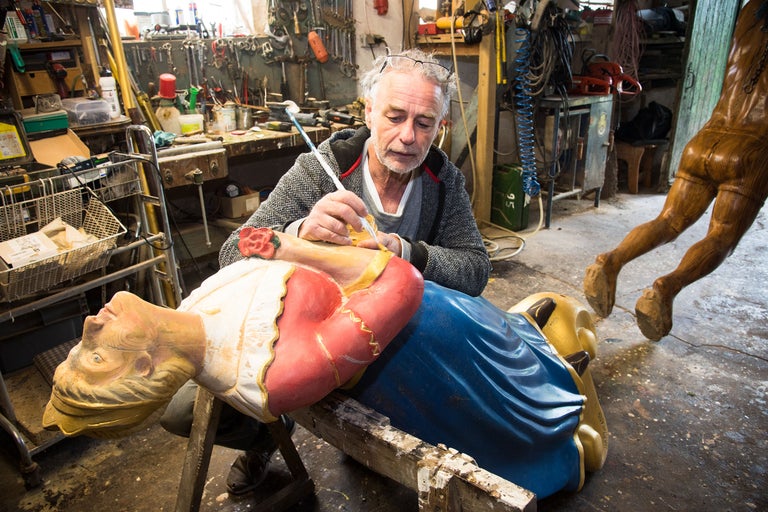 Ein Handwerker restauriert eine Holzfigur mit einem Pinsel in einer Werkstatt.