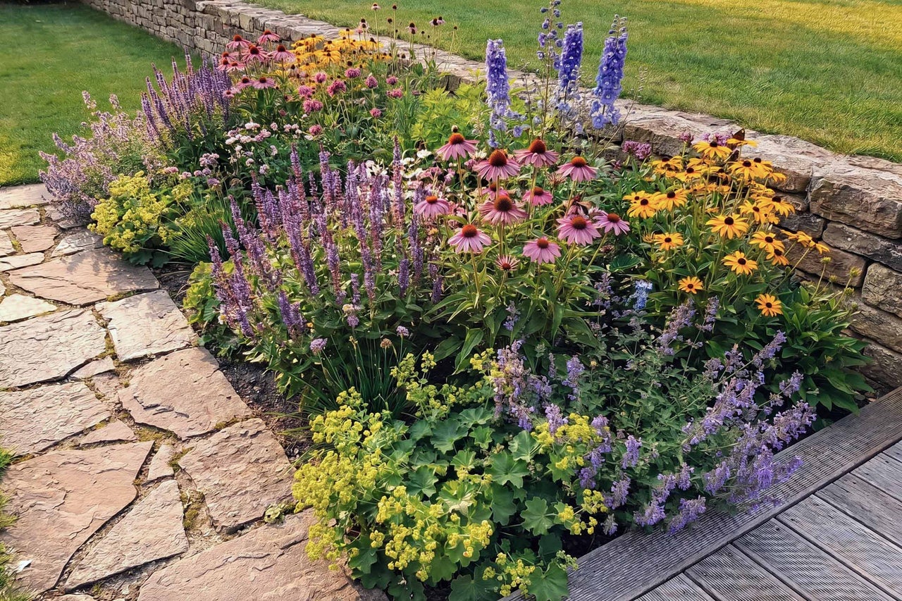Gartenbeet mit verschiedenen Stauden wie Sonnenhut, Salbei und Rittersporn neben einer Natursteinmauer und einem Steinpfad.