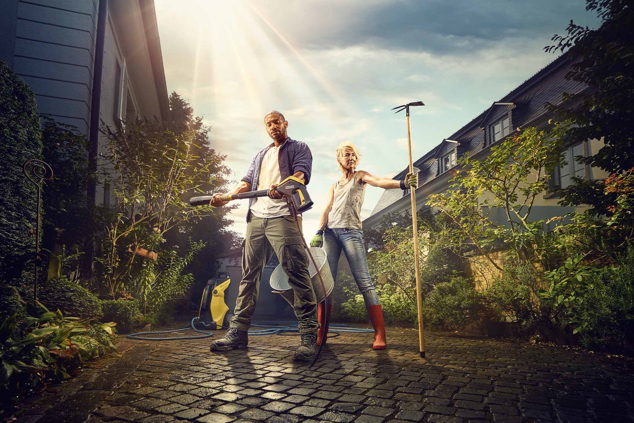 Ein Mann mit Hochdruckreiniger und eine Frau mit einer Hacke reinigen einen Hof mit Haus und Garten