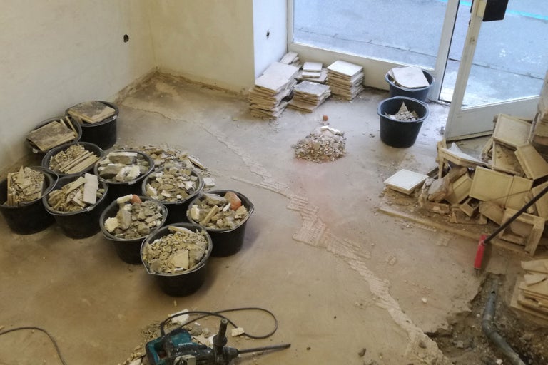 Renovierungsarbeiten in einem Raum mit Eimern voller Bauschutt, einem Bohrhammer und gestapelten Fliesen.