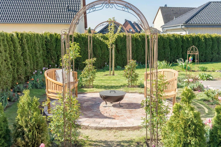 Gartenszene mit Rosenbogen, Sitzbänken und Feuerschale auf gepflastertem Untergrund