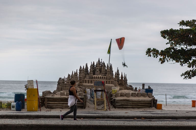 Eine komplexe Sandburg steht am Strand, mit einer Frau, die vorbeigeht, und einem Gleitschirmflieger im Hintergrund.