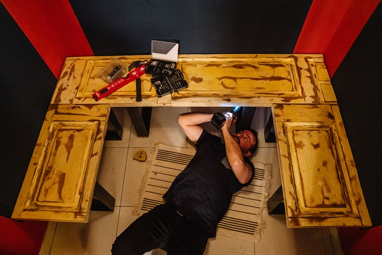 Mann liegt auf einem Teppich und montiert ein Möbelstück mit einem Akkuschrauber, Werkzeuge liegen bereit.