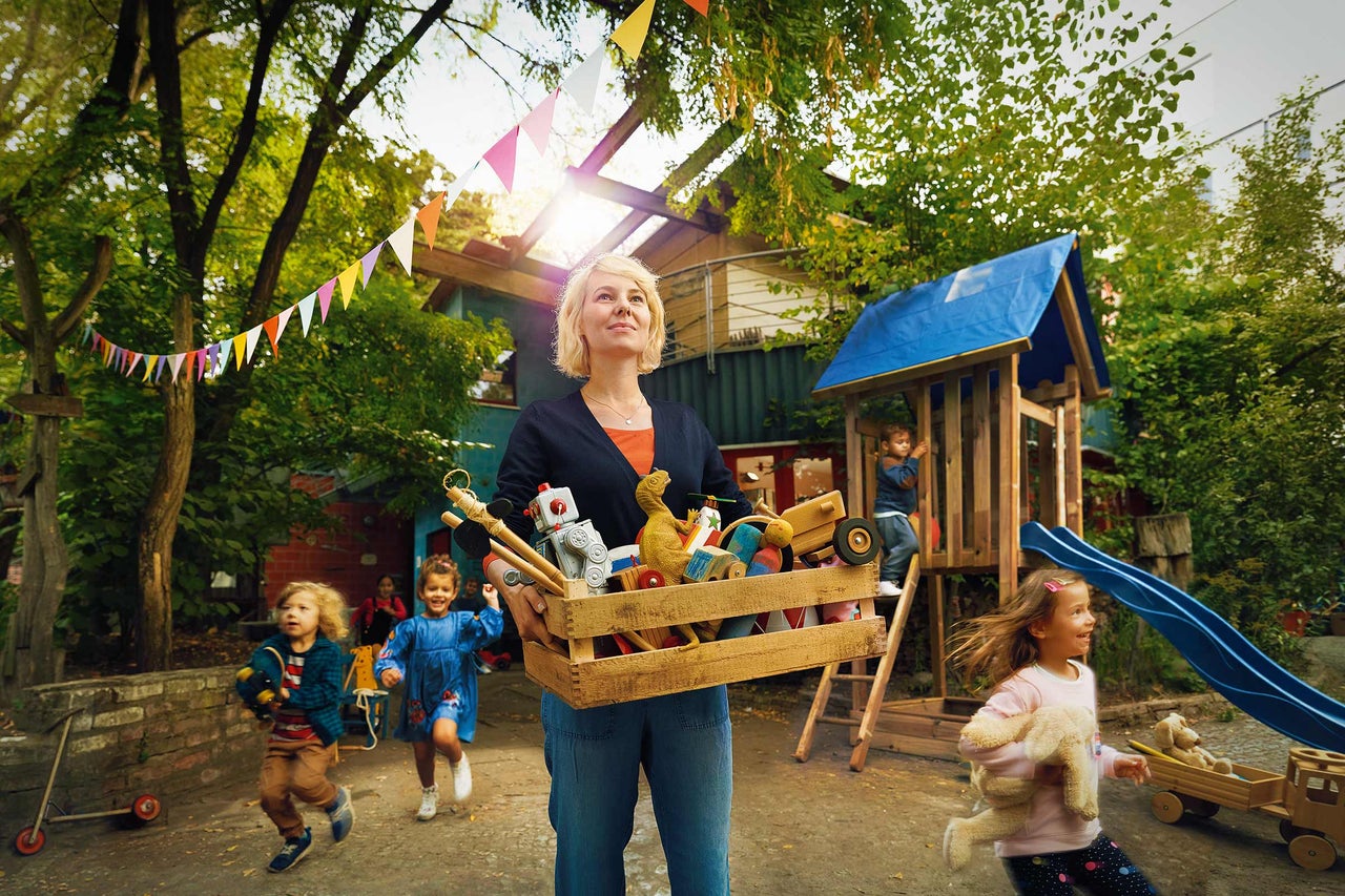 Eine Frau trägt eine Holzkiste voller Spielzeug vor einem Spielplatz mit Kindern