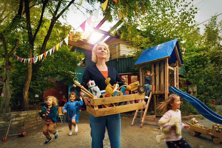 Eine Frau trägt eine Holzkiste voller Spielzeug vor einem Spielplatz mit Kindern