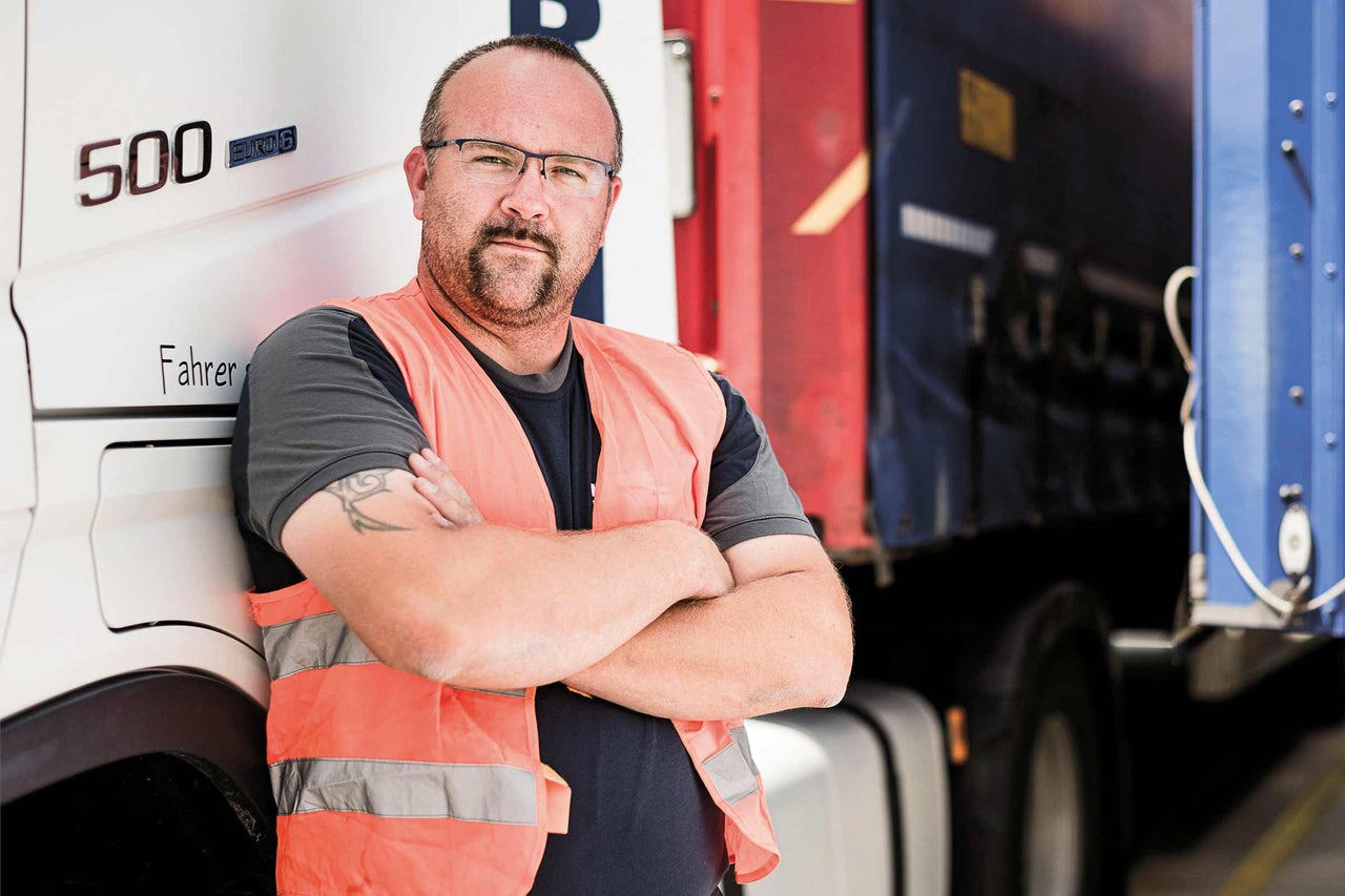 Ein Mann mit Brille und Warnweste lehnt an einem Lastkraftwagen.