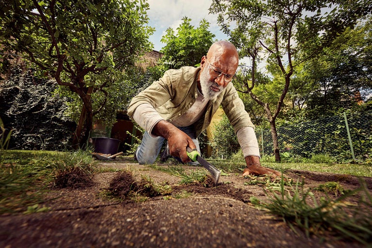 Ein Mann kniet im Garten und verwendet ein Handgartenwerkzeug zur Rasenpflege.