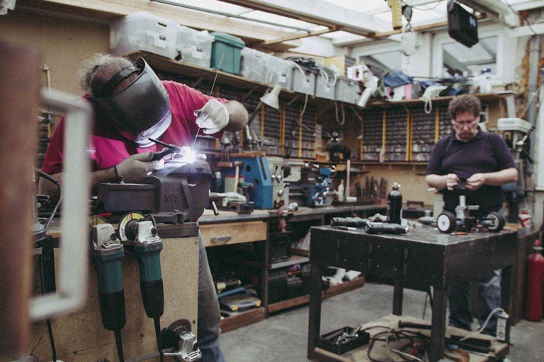 Werkstatt mit Schweißer, Werkbank, Werkzeugen und Regalen.
