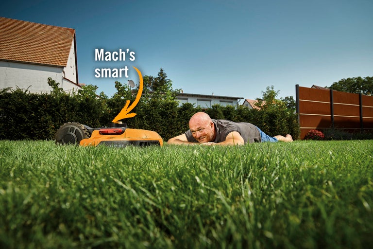 Ein Mann liegt im Gras neben einem Rasenmähroboter auf einer Rasenfläche.