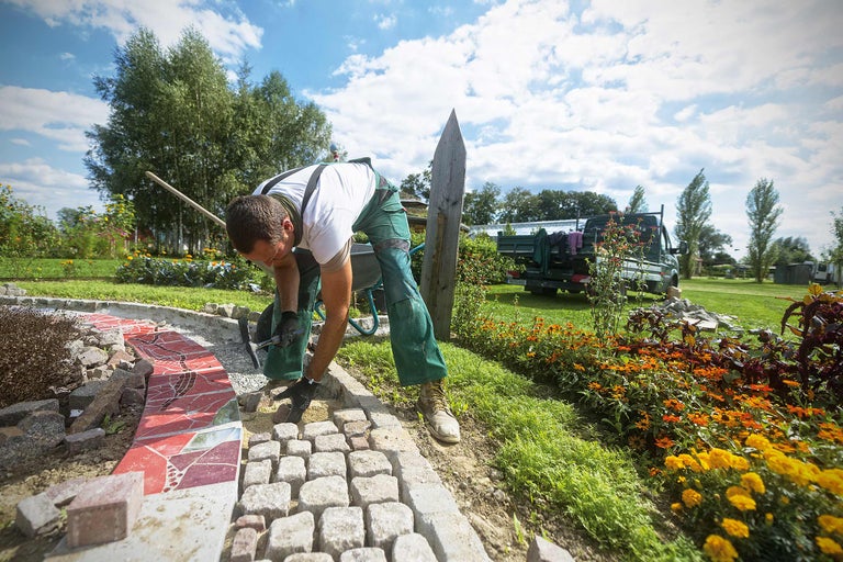 Ein Mann verlegt Pflastersteine in einem Garten mit Blumen und einem Fahrzeug im Hintergrund.