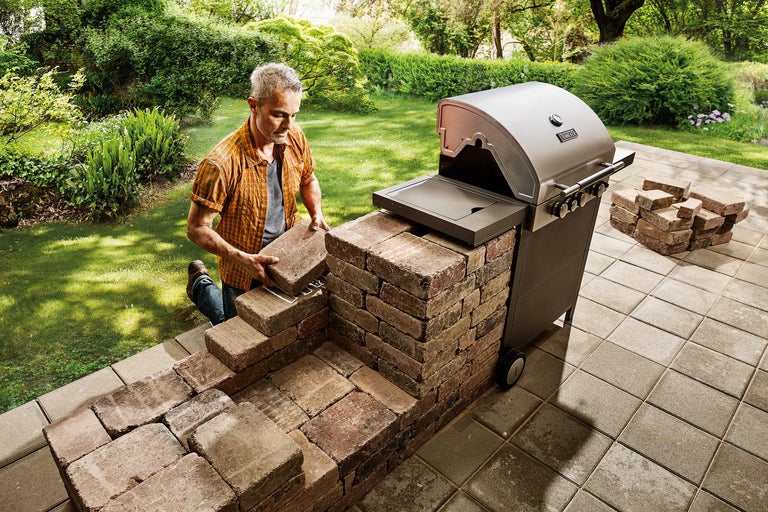 Mann baut eine Grillstation mit Steinen neben einem Gasgrill im Garten.