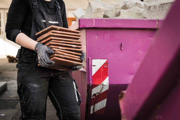 Eine Person trägt Dachziegel zu einem Bauschuttcontainer.
