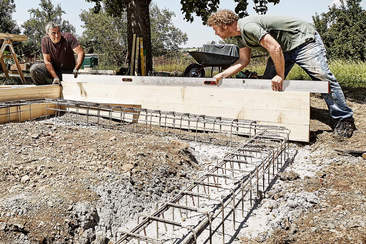 Zwei Männer arbeiten an einem Fundament mit Holzschalung und Stahlarmierung auf einer Baustelle.