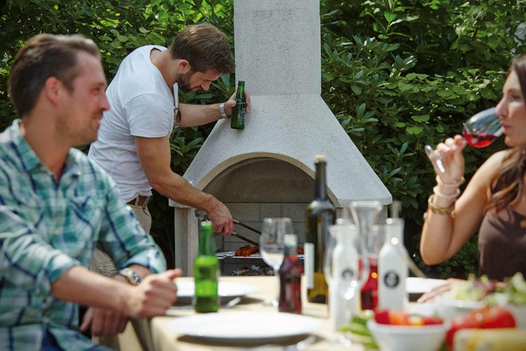 Personen grillen im Garten mit einem Gartengrill aus Stein und genießen Essen und Getränke an einem Tisch.