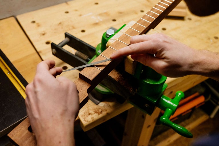 Eine Person bearbeitet mit einer Feile ein Gitarrenhals-Griffbrett, das in einem Schraubstock eingespannt ist.