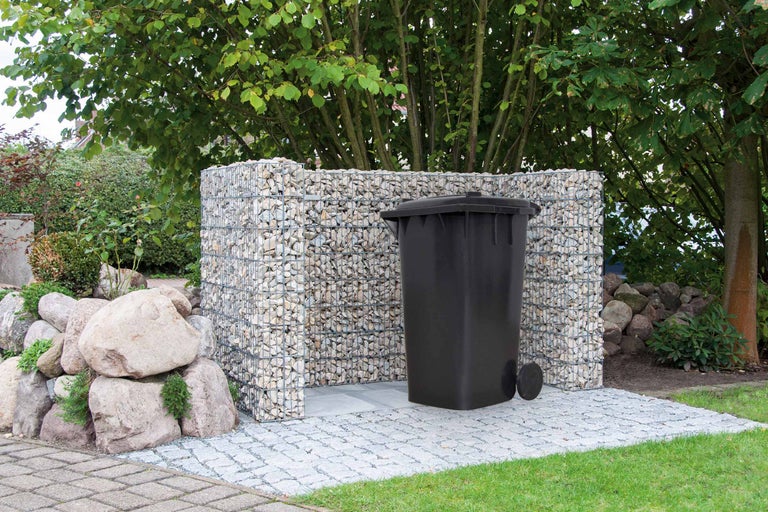 Eine Mülltonne steht vor einer Gabionenwand im Garten.